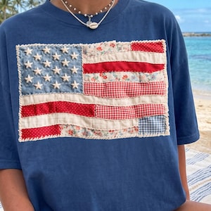 May include: A navy blue short-sleeve t-shirt featuring a rectangular patch on the chest designed to resemble the American flag. The flag patch is constructed from various fabric scraps in red, white, and blue, including floral and gingham patterns.