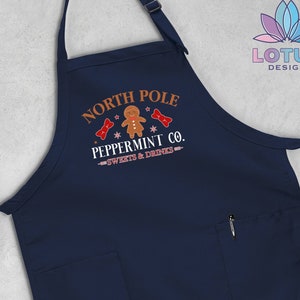 May include: A navy blue apron with a pocket and adjustable straps. The apron has a white design that says "North Pole Peppermint Co. - Sweets & Drinks" with a gingerbread man graphic.