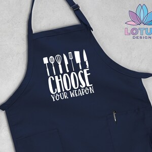 May include: A navy blue kitchen apron with a white pocket and a white graphic that says "Choose Your Weapon" with a row of kitchen utensils above the text.