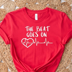 May include: Red t-shirt with white text that reads "The Beat Goes On" and a white heart with a heartbeat line.