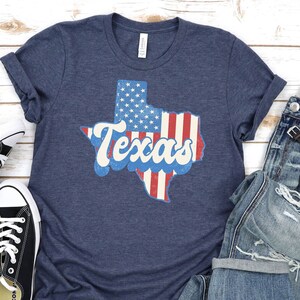 May include: A navy blue t-shirt with a distressed Texas graphic in red, white, and blue, featuring the state outline filled with the American flag design and the word "Texas". The shirt is paired with blue jeans and black sneakers.