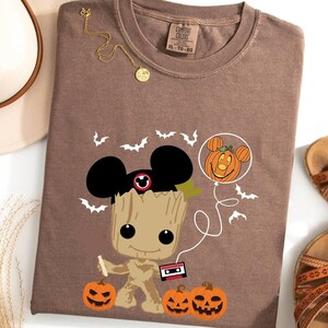 May include: Brown t-shirt with a cartoon image of a Groot character wearing Mickey Mouse ears and holding a pumpkin. The character is holding a cassette tape and a pumpkin. There are bats and a pumpkin balloon in the background.