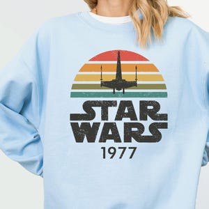 May include: Light blue sweatshirt featuring a retro Star Wars design. The graphic includes a silhouette of a spaceship against a sunset with the words "STAR WARS 1977" in bold, black letters. The sweatshirt is a solid light blue color.