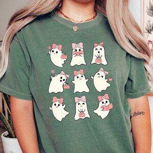 Może przedstawiać: Zielony T-shirt z grafiką przedstawiającą dziewięć białych duch&oacute;w w r&oacute;żowych kokardkach. Każdy duch wykonuje inną czynność, np. trzyma dynię, czyta książkę lub trzyma kwiat.