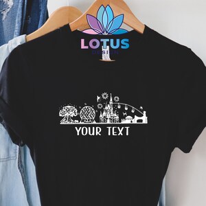 K&ouml;nnte beinhalten: Schwarzes T-Shirt mit einer wei&szlig;en Silhouette einer Themenpark-Skyline, einschlie&szlig;lich eines Schlosses, eines Riesenrads und eines Baumes. Der Text "YOUR TEXT" befindet sich unterhalb der Skyline.