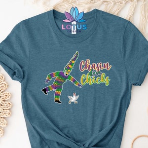 May include: A teal blue t-shirt with a graphic of a person in a Mardi Gras costume chasing a white chicken. The text "chasin' chicks" is printed on the shirt.