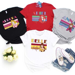 May include: Four different t-shirts featuring Disney princesses with retro-style graphics. The shirts are black, red, grey, and white. Each shirt features a different princess: Aurora, Ariel, Anna, and Belle. The shirts have a vintage look with colourful stripes and the princess's name in bold letters.
