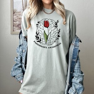 May include: A light green t-shirt featuring a black and white outline of a brain with a red tulip growing out of it. The text "Parkinson's Awareness" is printed below the brain.
