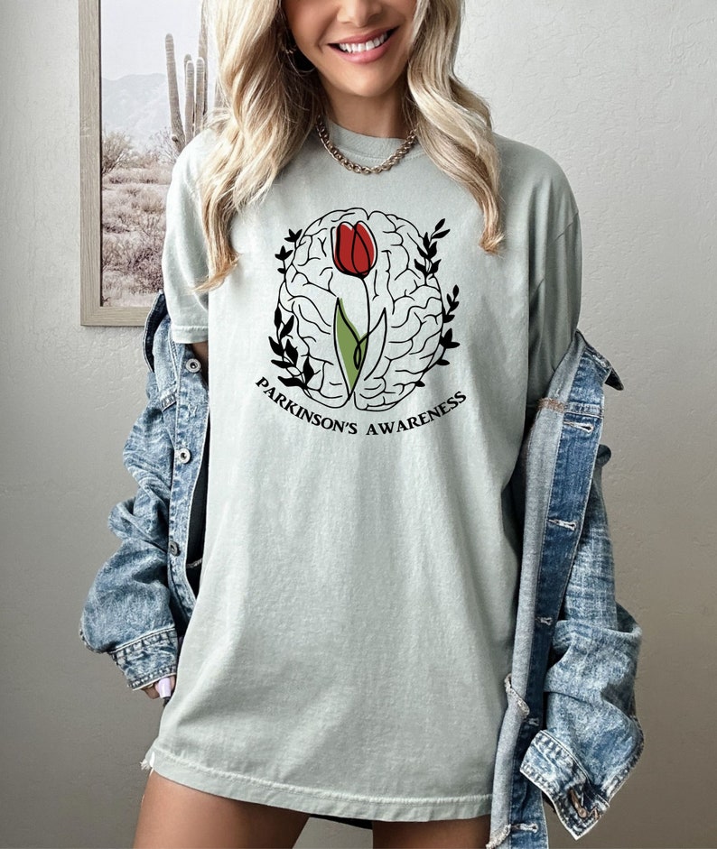 May include: A light green t-shirt featuring a black and white outline of a brain with a red tulip growing out of it. The text "Parkinson's Awareness" is printed below the brain.