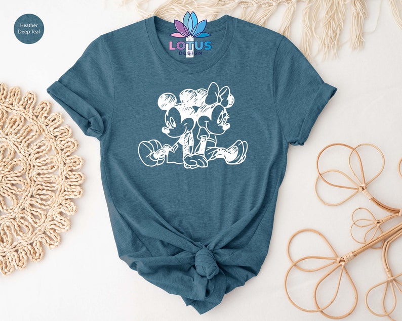 Pode incluir: Uma camiseta azul-petr&oacute;leo heather com um contorno branco do Mickey Mouse e da Minnie Mouse de m&atilde;os dadas. O texto "LOTUS DESIGN" est&aacute; impresso na camiseta.