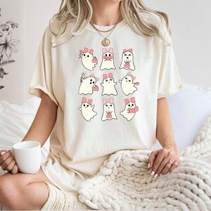 Może przedstawiać: Biały T-shirt z grafiką dziewięciu uroczych duch&oacute;w w r&oacute;żowych kokardkach. Duchy są w r&oacute;żnych pozach, niekt&oacute;re trzymają serca, niekt&oacute;re kwiaty, a niekt&oacute;re filiżanki kawy.