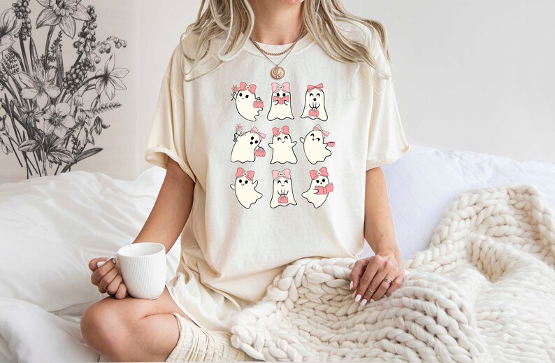 Może przedstawiać: Biały T-shirt z grafiką dziewięciu uroczych duch&oacute;w w r&oacute;żowych kokardkach. Duchy są w r&oacute;żnych pozach, niekt&oacute;re trzymają serca, niekt&oacute;re kwiaty, a niekt&oacute;re filiżanki kawy.