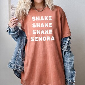 May include: A rust-colored oversized t-shirt with the text "Shake Shake Shake Senora" printed in white.