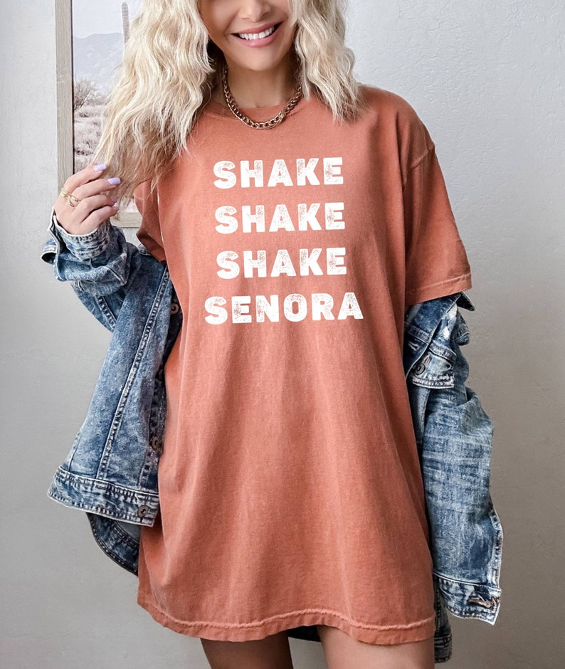 May include: A rust-colored oversized t-shirt with the text "Shake Shake Shake Senora" printed in white.