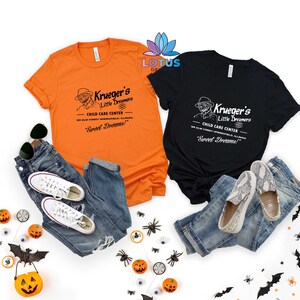 May include: Two t-shirts with a vintage-style graphic of a child care center called "Krueger's Little Dreamers". The text on the shirts reads "Child Care Center" and "Sweet Dreams!" The shirts are orange and black and are laid out on a white background with Halloween decorations.