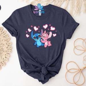 K&ouml;nnte beinhalten: Ein T-Shirt in Marineblau mit einem Aufdruck von Stitch und Angel aus Lilo & Stitch. Der Aufdruck zeigt Stitch und Angel, die sich umarmen, mit Herzen um sie herum.