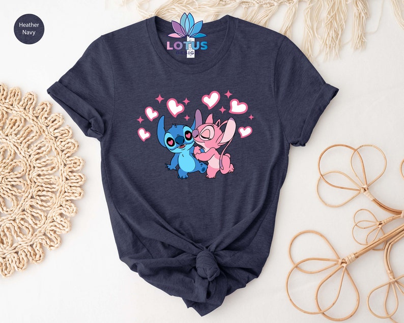 K&ouml;nnte beinhalten: Ein T-Shirt in Marineblau mit einem Aufdruck von Stitch und Angel aus Lilo & Stitch. Der Aufdruck zeigt Stitch und Angel, die sich umarmen, mit Herzen um sie herum.