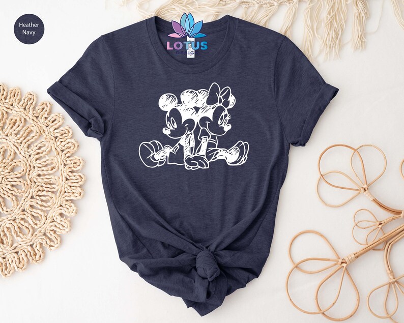Pode incluir: Uma camiseta azul marinho heather com um gr&aacute;fico branco do Mickey Mouse e da Minnie Mouse sentados juntos. O gr&aacute;fico &eacute; um estilo de desenho de linha simples.