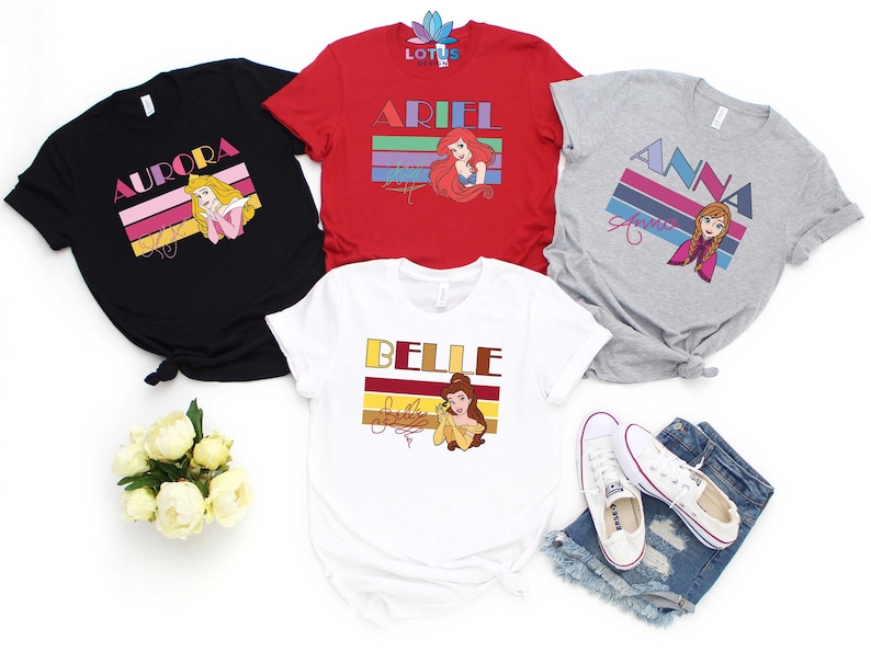 May include: Four different t-shirts featuring Disney princesses with retro-style graphics. The shirts are black, red, grey, and white. Each shirt features a different princess: Aurora, Ariel, Anna, and Belle. The shirts have a vintage look with colourful stripes and the princess's name in bold letters.