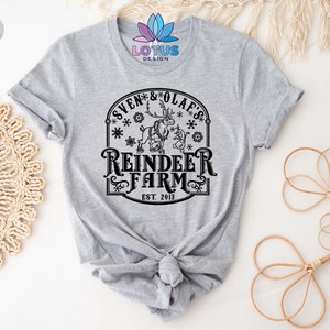 May include: A gray t-shirt with a black graphic design of a reindeer and a snowman. The design includes the text "Sven & Olaf's Reindeer Farm Est. 2013".