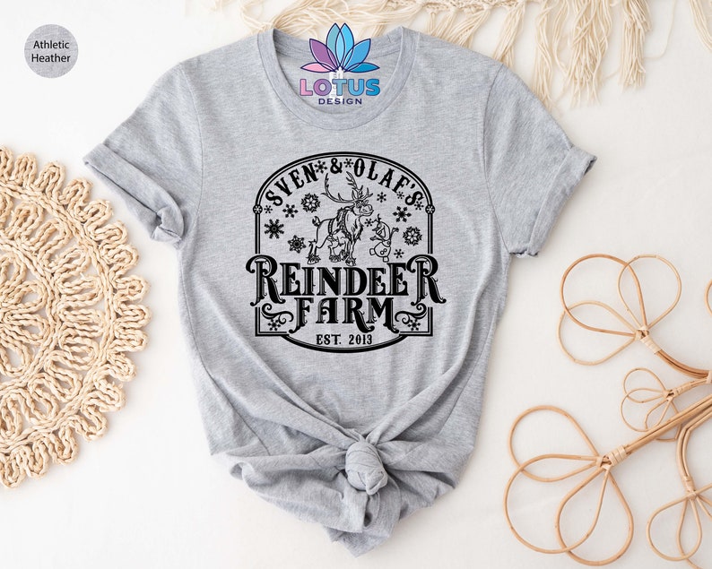 May include: A gray t-shirt with a black graphic design of a reindeer and a snowman. The design includes the text "Sven & Olaf's Reindeer Farm Est. 2013".