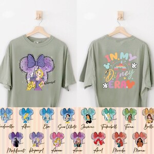 Puede incluir: Una camiseta verde con un dise&ntilde;o de orejas de Mickey Mouse en morado y blanco con una princesa de Disney. La parte posterior de la camiseta tiene el texto "In My Disney Era" en colores arco&iacute;ris. La camiseta se muestra en una percha con una variedad de otras camisetas con diferentes princesas de Disney.