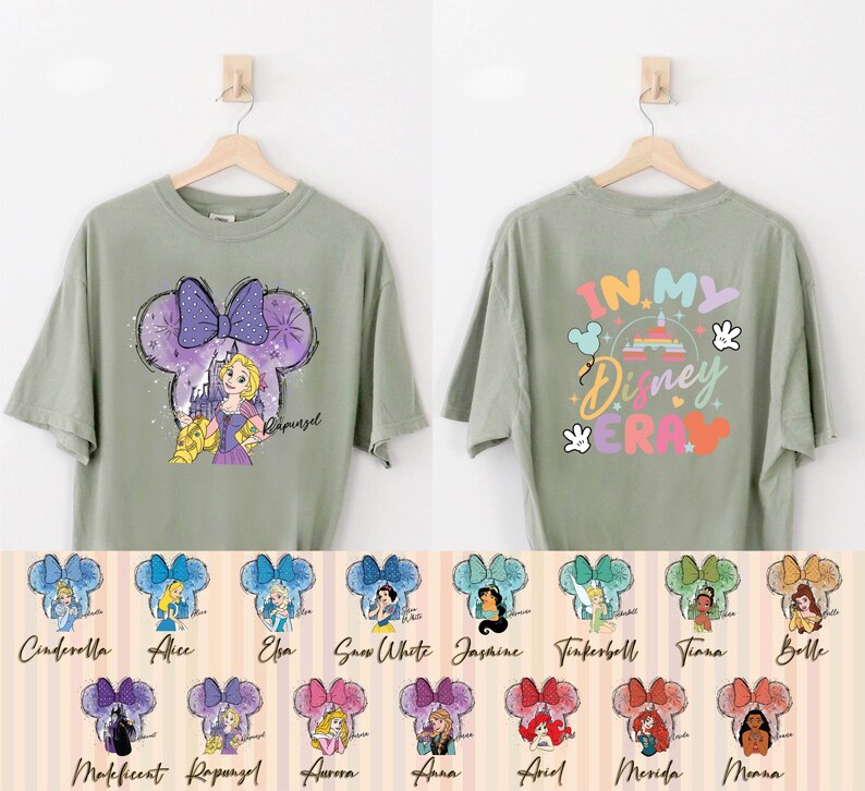Puede incluir: Una camiseta verde con un dise&ntilde;o de orejas de Mickey Mouse en morado y blanco con una princesa de Disney. La parte posterior de la camiseta tiene el texto "In My Disney Era" en colores arco&iacute;ris. La camiseta se muestra en una percha con una variedad de otras camisetas con diferentes princesas de Disney.