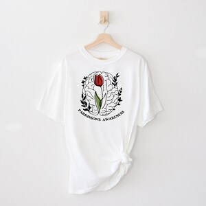 May include: White t-shirt featuring a black and white outline of a brain with a red tulip growing from it. The text "Parkinson's Awareness" is written around the brain.