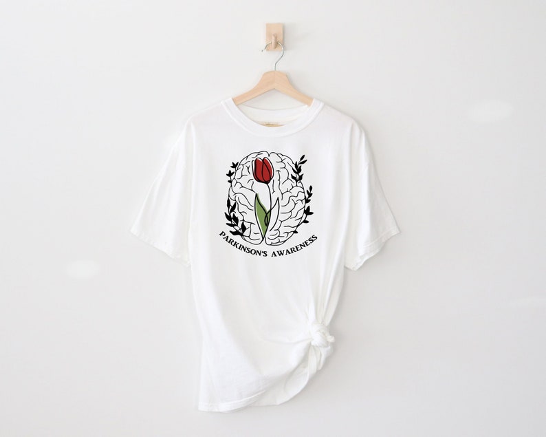 May include: White t-shirt featuring a black and white outline of a brain with a red tulip growing from it. The text "Parkinson's Awareness" is written around the brain.