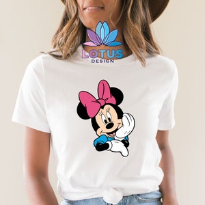 K&ouml;nnte beinhalten: Wei&szlig;es T-Shirt mit einem Cartoon-Bild von Minnie Mouse, die eine rosa Schleife tr&auml;gt. Der Text "LOTUS DESIGN" ist auf das Shirt gedruckt.