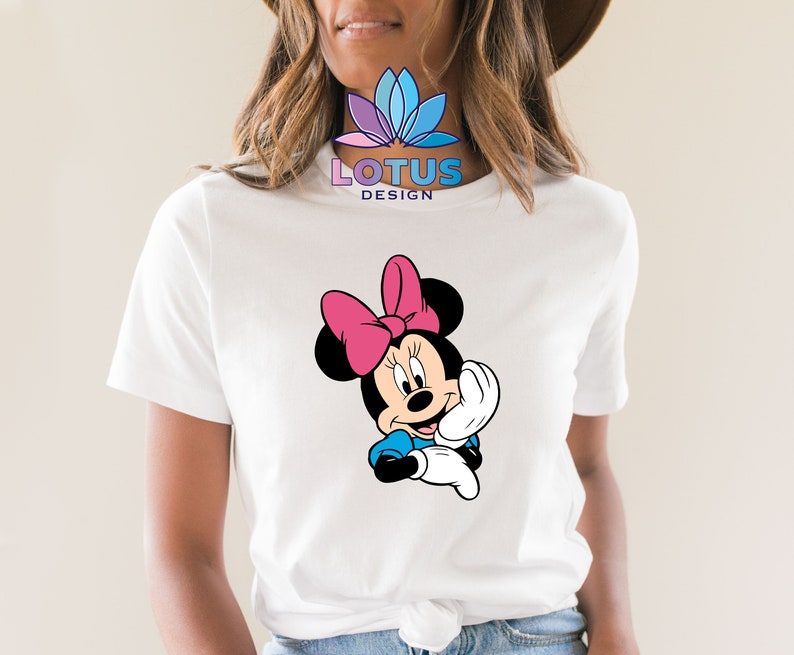 K&ouml;nnte beinhalten: Wei&szlig;es T-Shirt mit einem Cartoon-Bild von Minnie Mouse, die eine rosa Schleife tr&auml;gt. Der Text "LOTUS DESIGN" ist auf das Shirt gedruckt.