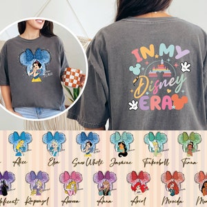 Puede incluir: Una camiseta gris con el texto "IN MY Disney ERA" en la espalda. La parte delantera de la camiseta presenta una imagen de dibujos animados de Blancanieves con un lazo azul y el texto "Snow White". Hay 12 personajes de princesas Disney diferentes en la parte inferior de la imagen, cada uno con un lazo de color diferente.
