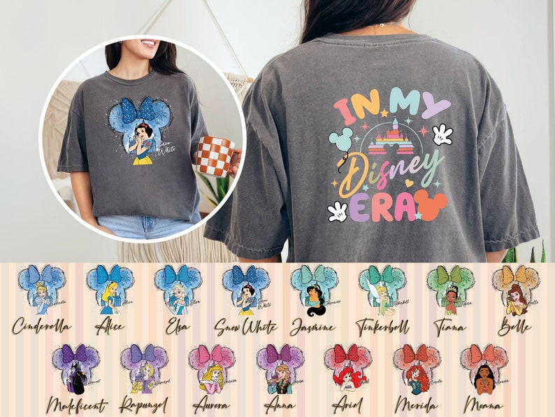 Puede incluir: Una camiseta gris con el texto "IN MY Disney ERA" en la espalda. La parte delantera de la camiseta presenta una imagen de dibujos animados de Blancanieves con un lazo azul y el texto "Snow White". Hay 12 personajes de princesas Disney diferentes en la parte inferior de la imagen, cada uno con un lazo de color diferente.
