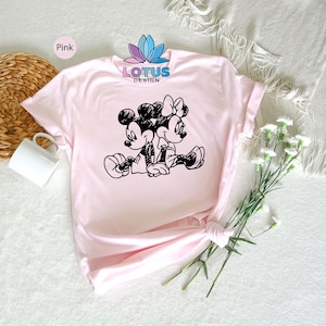 Pode incluir: Camiseta rosa com uma ilustra&ccedil;&atilde;o em preto e branco do Mickey Mouse e da Minnie Mouse de m&atilde;os dadas. A camiseta tem o texto "LOTUS DESIGN" impresso.