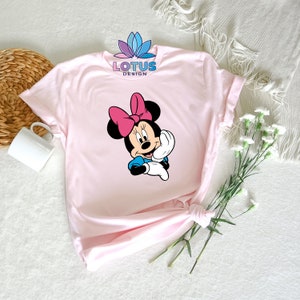 K&ouml;nnte beinhalten: Rosa T-Shirt mit einem Aufdruck von Minnie Mouse. Der Aufdruck zeigt Minnie Mouse mit einer rosa Schleife, einem blauen Kleid und wei&szlig;en Handschuhen. Der Text "LOTUS DESIGN" ist auf das T-Shirt gedruckt.