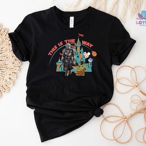 May include: Black t-shirt featuring a graphic of Star Wars characters, including Yoda and Mandalorian, in front of a castle. The text "This is the way" is printed on the shirt.
