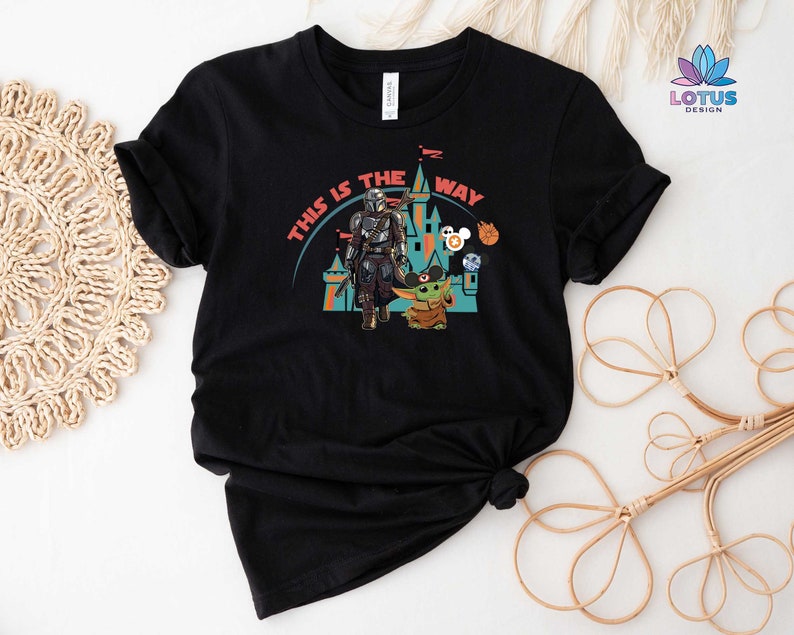 May include: Black t-shirt featuring a graphic of Star Wars characters, including Yoda and Mandalorian, in front of a castle. The text "This is the way" is printed on the shirt.