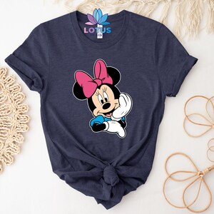 K&ouml;nnte beinhalten: Ein marineblaues T-Shirt mit einem Aufdruck von Minnie Maus. Minnie Maus tr&auml;gt eine rosa Schleife und ein blaues Kleid. Sie l&auml;chelt und winkt.