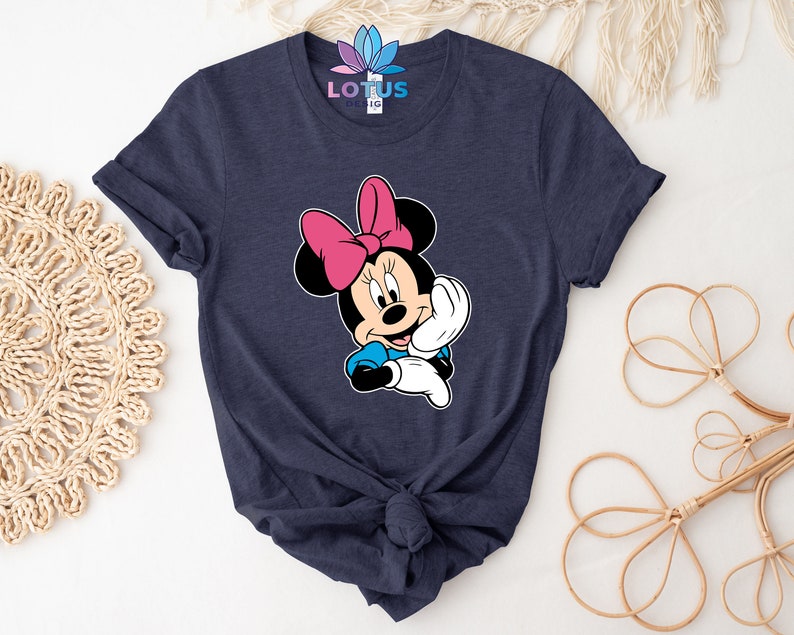 K&ouml;nnte beinhalten: Ein marineblaues T-Shirt mit einem Aufdruck von Minnie Maus. Minnie Maus tr&auml;gt eine rosa Schleife und ein blaues Kleid. Sie l&auml;chelt und winkt.