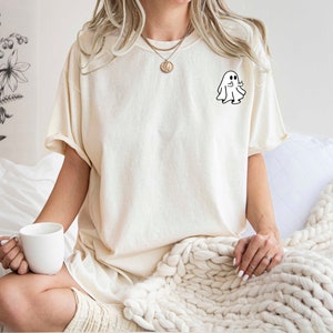 May include: A woman wearing a cream colored t-shirt with a small white ghost graphic on the left chest. She is sitting on a bed with a white blanket and holding a white mug.