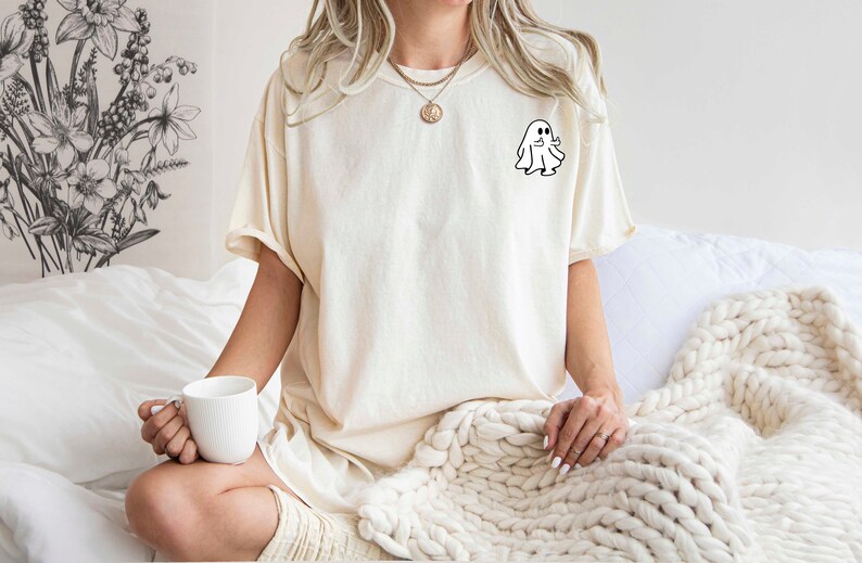 May include: A woman wearing a cream colored t-shirt with a small white ghost graphic on the left chest. She is sitting on a bed with a white blanket and holding a white mug.