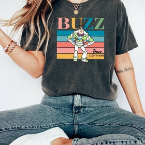 May include: A dark gray t-shirt with a retro-style graphic of Buzz Lightyear from Toy Story. The graphic features a colorful striped background and the text "Buzz Lightyear".