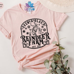May include: A pink t-shirt with a black graphic design featuring a reindeer and a snowman with the text "Sven & Olaf's Reindeer Farm Est. 2013".
