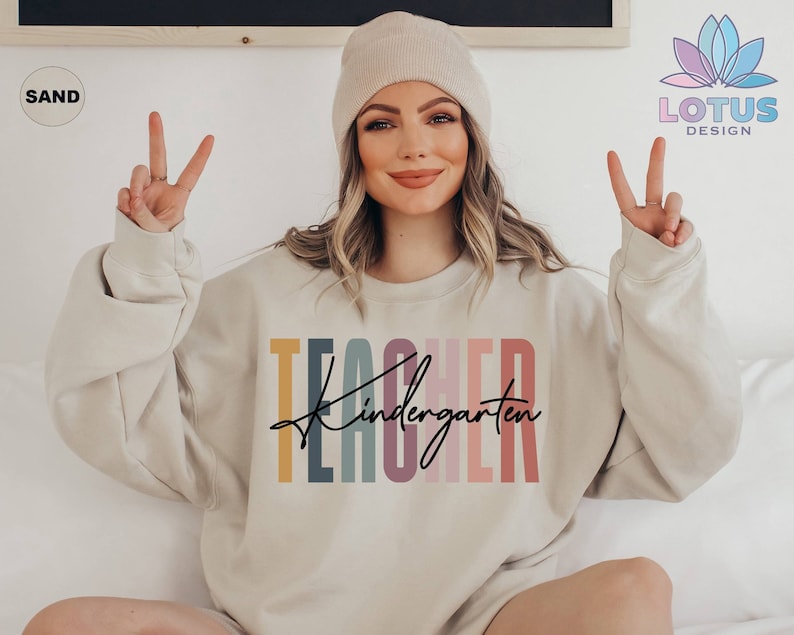 K&ouml;nnte beinhalten: Ein hellbeiger Pullover mit dem Text "TEACHER kindergarten" in bunten vertikalen Streifen. Der Text ist in einer modernen, handschriftlichen Schrift.