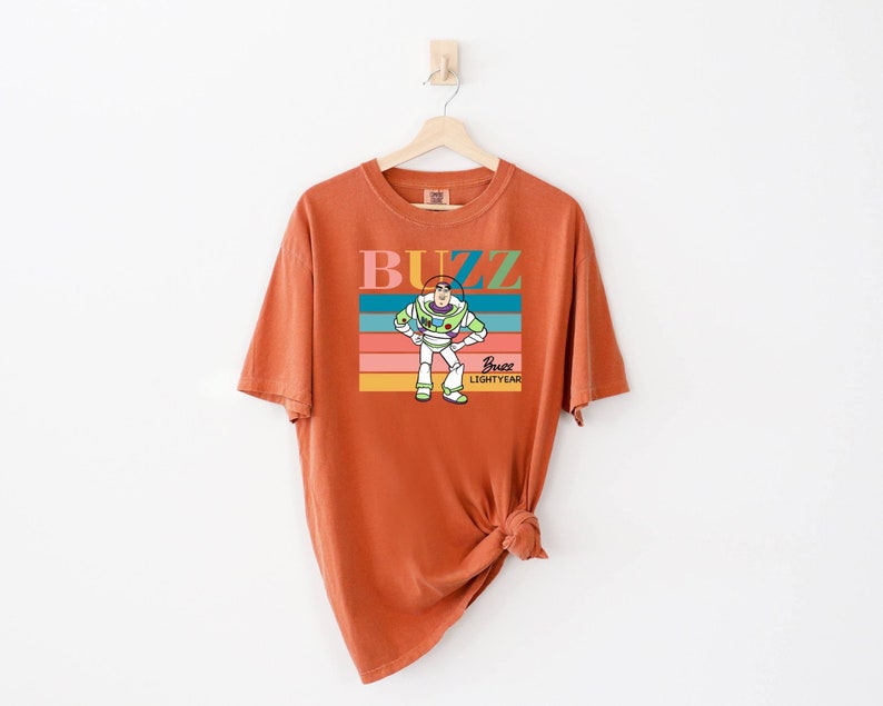 May include: Orange t-shirt with a retro-style graphic of Buzz Lightyear from Toy Story. The graphic features a rainbow stripe background with the text "Buzz Lightyear" and a cartoon image of Buzz Lightyear.