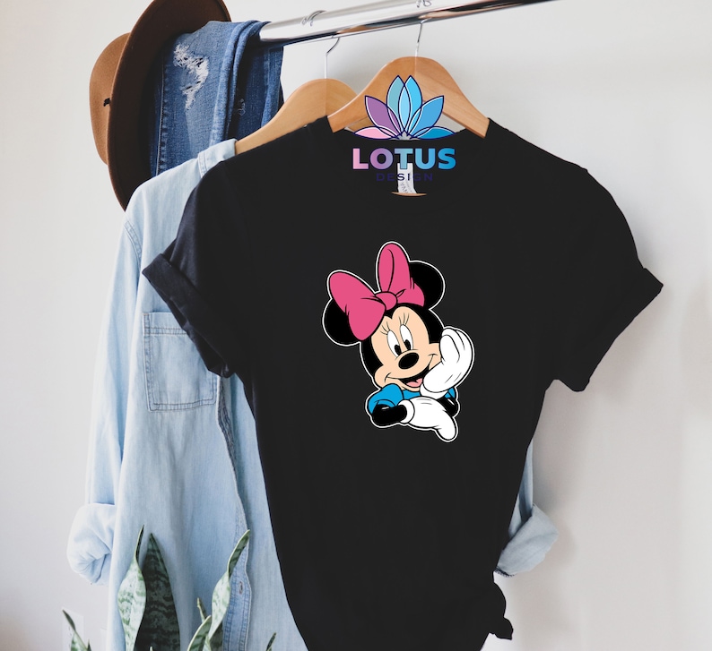 K&ouml;nnte beinhalten: Schwarzes T-Shirt mit einem Cartoon-Bild von Minnie Mouse, die eine rosa Schleife tr&auml;gt. Der Text "LOTUS DESIGN" ist auf dem Shirt aufgedruckt.