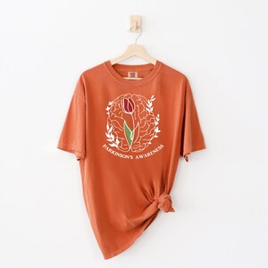 May include: A rust-coloured t-shirt with a white outline of a brain with a tulip growing out of it. The text "Parkinson's Awareness" is written below the brain.