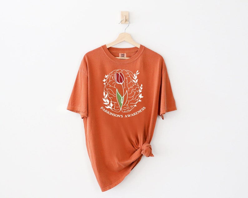 May include: A rust-coloured t-shirt with a white outline of a brain with a tulip growing out of it. The text "Parkinson's Awareness" is written below the brain.
