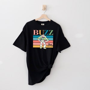 May include: Black t-shirt with a retro-style graphic of Buzz Lightyear from Toy Story. The graphic features Buzz in his space ranger suit with the text "Buzz Lightyear" and a rainbow stripe background.