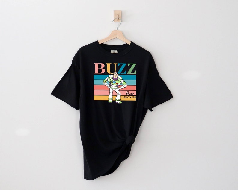 May include: Black t-shirt with a retro-style graphic of Buzz Lightyear from Toy Story. The graphic features Buzz in his space ranger suit with the text "Buzz Lightyear" and a rainbow stripe background.